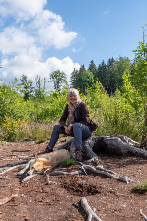 Frau sitzt auf einem Baumstumpf und ein Hund liegt ihr zu Füßen