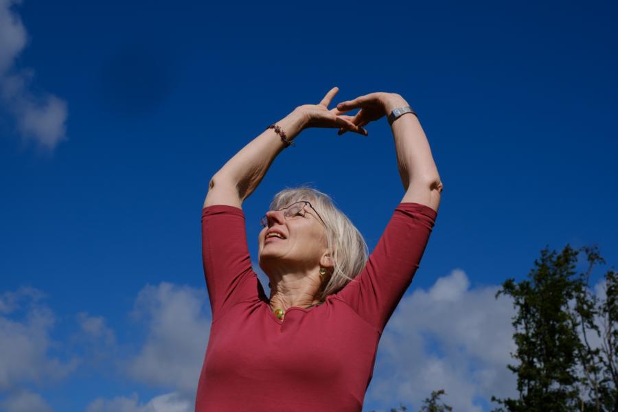 Frau mit über den Kopf erhobenen Händen, die sich an den Fingern berühren. Vor strahlend blauem Himmel.